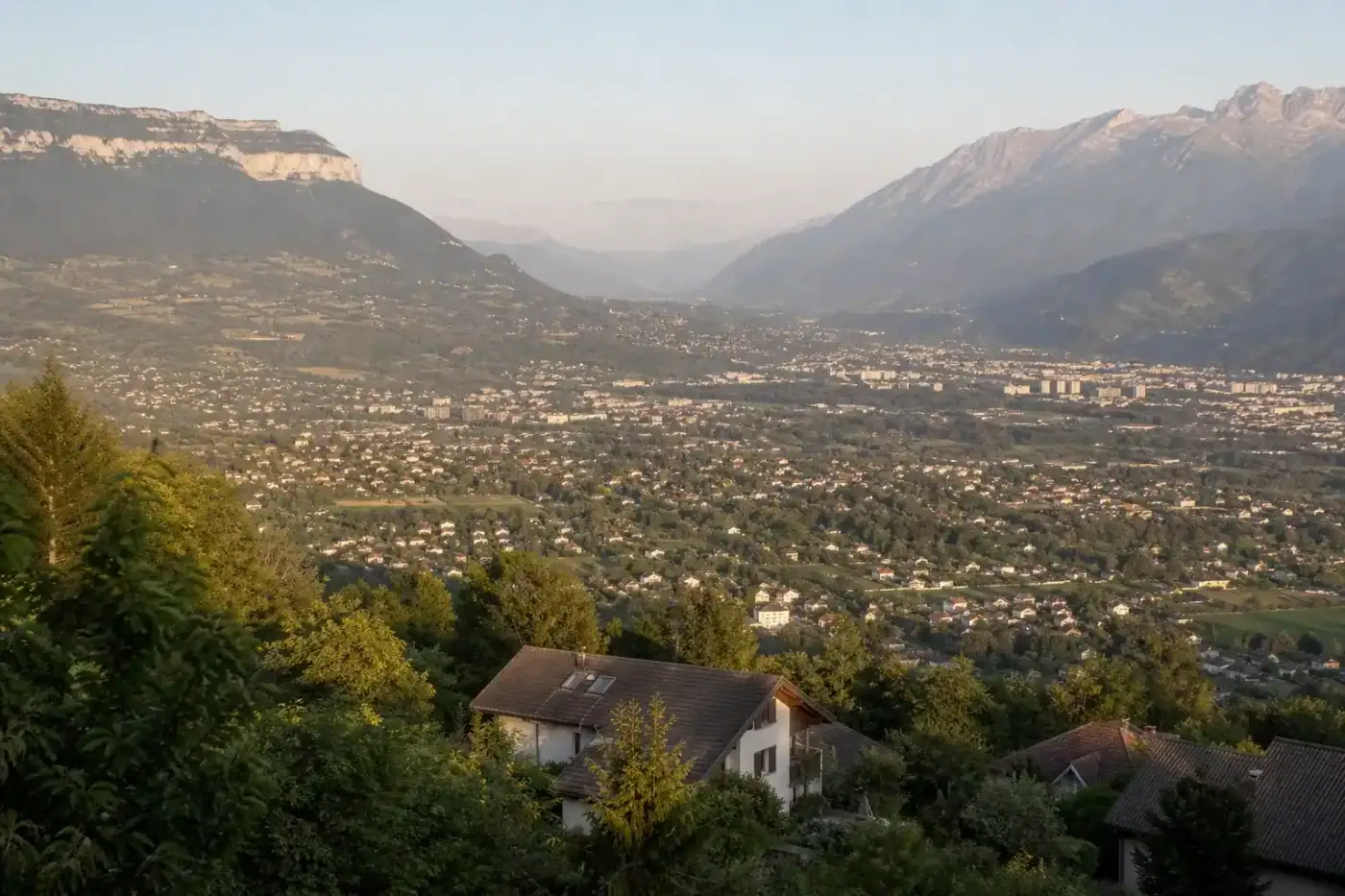 vue large de l'agglomération grenobloise depuis les hauteurs, maisons individuelles et lotissements en fond de vallée, massifs du Vercors et de Belledonne autour, lumière de fin de journée, aucune personne visible